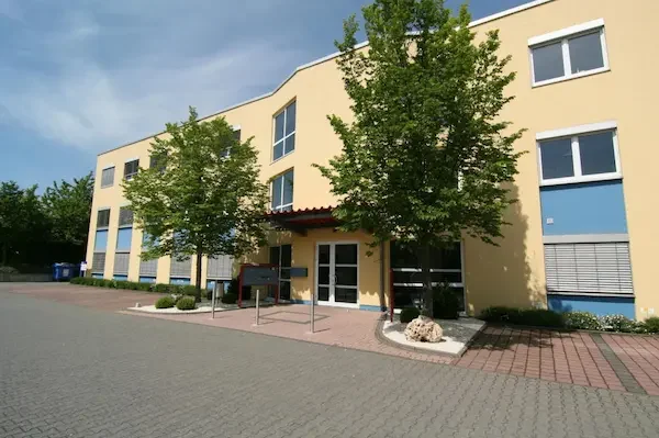 Büro Gelbes Geschäftsgebäude mit Bäumen und Parkplatz. Eingangstüren sind sichtbar.
